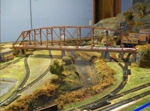 Red steel bridge over train tracks and water on the model railroad layout. 