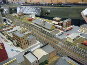 Model train tracks between houses and buildings with cars and trucks.