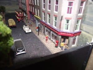 People walking along the sidewalk on the model railroad with cars.