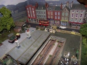 Storefronts and vehicles on the model train layout with background scenery. 