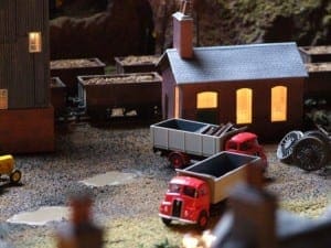 Two red dump trucks parked outside a red house with a chimney on the model train layout. 