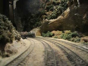 Two model trains run through the train tracks between mountains covered with trees.