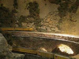 Yellow model train running through track beside mountain rubble.