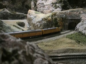 yellow model train entering a tunnel surrounded by mountains