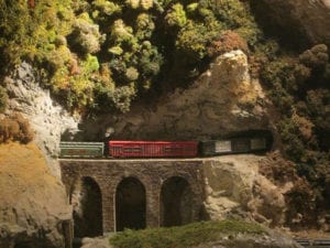 Model train exiting a tunnel and crossing a bridge located underneath plenty of trees. 