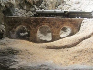 Model train crossing an arch bridge in between mountains on the model railroad layout.