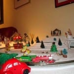 Ground-level view of the Christmas-themed railroad with a cartoon character, a red car carrying a Christmas tree, and the model train. 