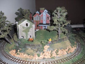 two people leaning on the fence surrounded by model train tracks