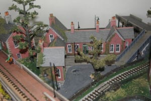 woman sitting on bench near the model railroad tracks. behind her are houses and a large tree.