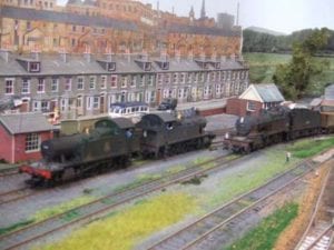 Two weathered model trains passing by a row of houses on the model railroad.
