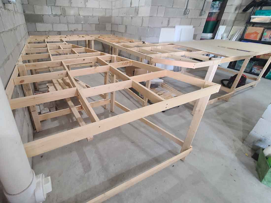 Large L-shaped model railroad benchwork frame in a basement with open grid joists and partial plywood top