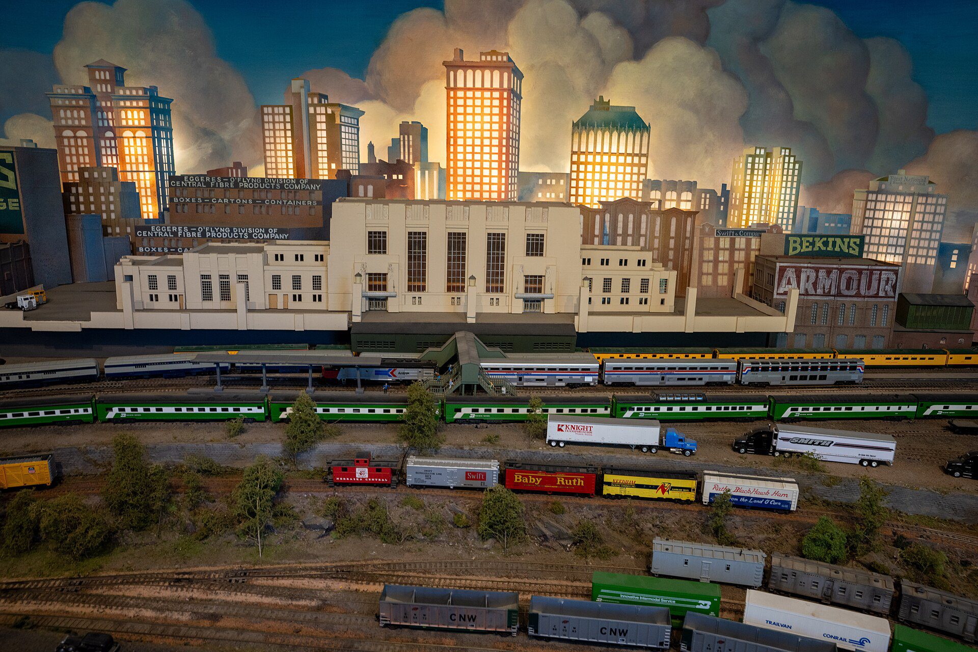 HO model railroad city scene with lit skyscrapers, a station, a passenger train and a freight train in the foreground