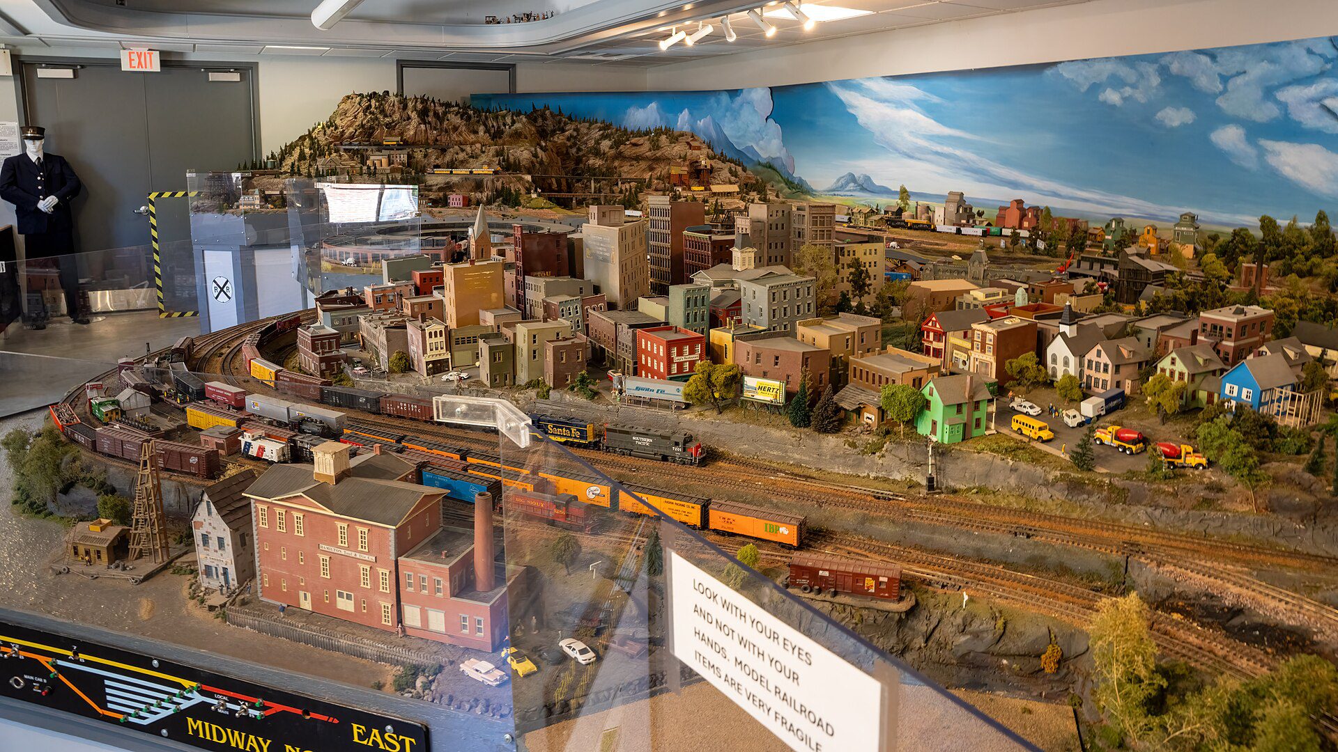 A large HO model railroad with a town, a rocky hill, a painted sky and freight trains running through the yard